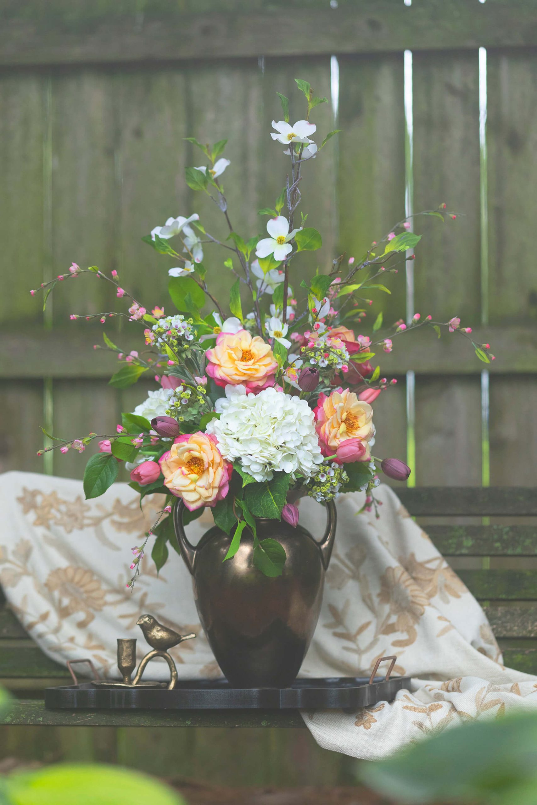 Luxury permanent botanical arrangement with peach roses, white hydrangeas, and tall branches in a bronze urn vase on a tray.